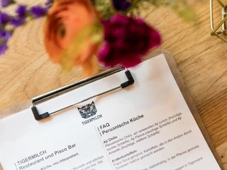 Tigermilch Das Bild zeigt im Vordergrund eine Speisekarte des Restaurants Tigermilch, auf einem Klemmbrett auf einem Holztisch, mit unscharfen Blüten im Hintergrund.The image shows, in the foreground, a menu of the restaurant Tigermilch displayed on a clipboard resting on a wooden table, with blurred flowers in the background.