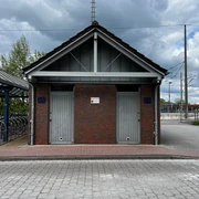 Öffentliche Toilette am Bahnhof.jpg Modernes, symmetrisches Ziegelgebäude am Bahnhof mit zwei getrennten Eingängen und asphaltiertem Vorplatz.