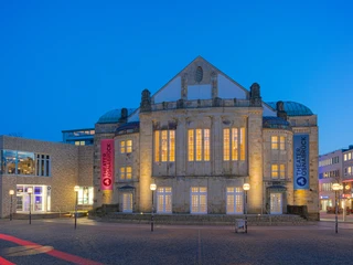 Historisches Theatergebäude in abendlicher Beleuchtung, flankiert von modernen Strukturen und Banner.