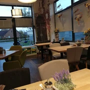 Bäckerei Ripken_Edeka_Augustfehn_2022.jpg Das Innere der Bäckerei Ripken in Augustfehn strahlt eine gemütliche und einladende Atmosphäre aus. Holzvertäfelte Möbel und dezente Dekorationen, wie Trockenblumen und moderne Lampen, schaffen ein stilvolles, rustikales Ambiente. Große Fenster sorgen für viel Tageslicht und geben den Blick auf die Umgebung frei, während die Tischdekoration mit kleinen Kräutertöpfen den ländlichen Charme unterstreicht.