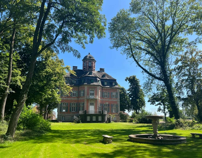 schloss-arff-eventlocation-park-feiern.jpg Rasenfläche mit Bäumen und Springbrunnen, im Hintergrund steht das Schloss ArffLawn with trees and fountain, with Arff Castle in the background