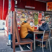 Kebapland Zwei Männer sitzen vor dem Imbiss an einem Holztisch und essen Kebap Dürüm. Die Fassade ist rot gestrichen und die Fenster mit Stickern beklebt.Two men are sitting at a wooden table in front of the snack bar eating kebap dürüm. The façade is painted red and the windows are covered with stickers.