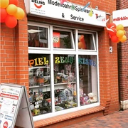 Ansicht.png Ein rotes Backsteingebäude mit einem Schaufenster voller Spielzeug und bunten Luftballons vorne.