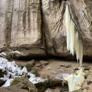 Gautschgrotte Gefrorener Wasserfall hängt von einer steinigen Klippe, umgeben von schneebedeckten Bäumen und Felsen in einer winterlichen Landschaft.