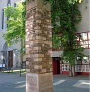 Schmitz column Ein hoher, mit unterschiedlich farbigen Steinen verkleideter Pfeiler steht auf einem gepflasterten Platz vor einer Kirche. Umgeben von Bäumen und einem Gebäude mit roten Fensterrahmen.A tall pillar clad with differently colored stones stands on a paved square in front of a church. It is surrounded by trees and a building with red window frames.