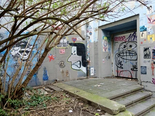 Wall of Fame Im Bild ist ein Eingang zu sehen, der mit farbenfrohen Graffiti bedeckt ist. Der Bereich ist von einem Baum umgeben, dessen Äste sich über den gepflasterten Boden erstrecken.The image shows an entrance covered with colorful graffiti. The area is surrounded by a tree, whose branches extend over the paved ground.