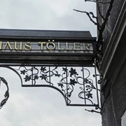 House Töller Ein kunstvoll geschmiedetes Schild mit "Haus Töller" hängt an einer Hauswand aus grauem Putz.An artistically forged sign with "Haus Töller" hangs on a gray plaster house wall.