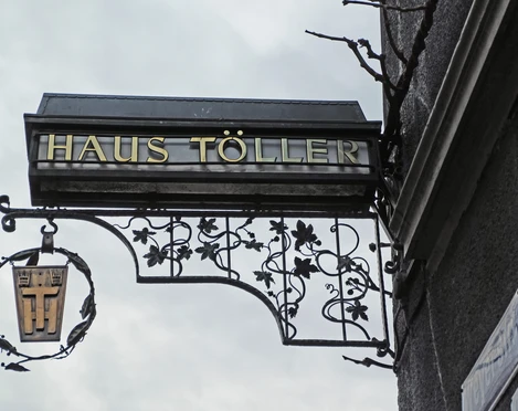 House Töller Ein kunstvoll geschmiedetes Schild mit "Haus Töller" hängt an einer Hauswand aus grauem Putz.An artistically forged sign with "Haus Töller" hangs on a gray plaster house wall.