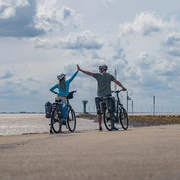 Zwei Radfahrer am Hafen heben die Hände zum High-Five, im Hintergrund erstreckt sich das Meer.