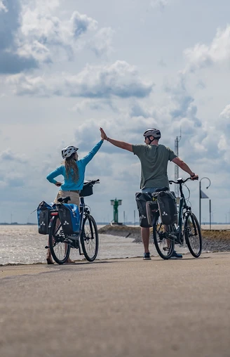 Emden-Außenhafen_01-CC_BY_SA-MaximilianSemsch_EmsRadweg.jpg Zwei Radfahrer am Hafen heben die Hände zum High-Five, im Hintergrund erstreckt sich das Meer.