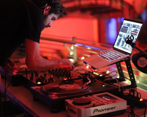 Liquid Sound Club Ein DJ performt in einem gedämpft beleuchteten Raum mit moderner Ausrüstung, umgeben von rotem Licht.