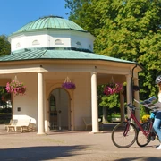 Ein Radfahrer mit Helm studiert eine Karte vor einem historischen Tempel mit Säulen und Blumenampeln.