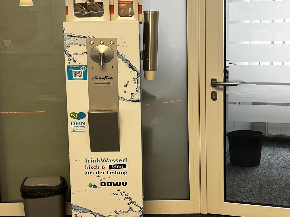 Trinkwasserspender im Rathaus_Manuela Baumhöfer.jpg Silberner Trinkwasserspender mit Bechern und Infomaterial im Eingangsbereich eines Büros.