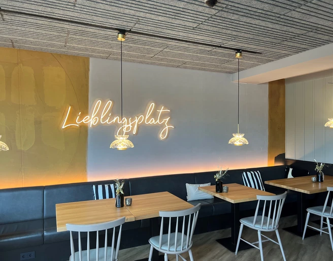 Thöles in Bücken.jpg Gemütlicher Gastraum mit Holz-Tischen, weißen Stühlen und neonleuchtendem Schriftzug „Lieblingsplatz“.