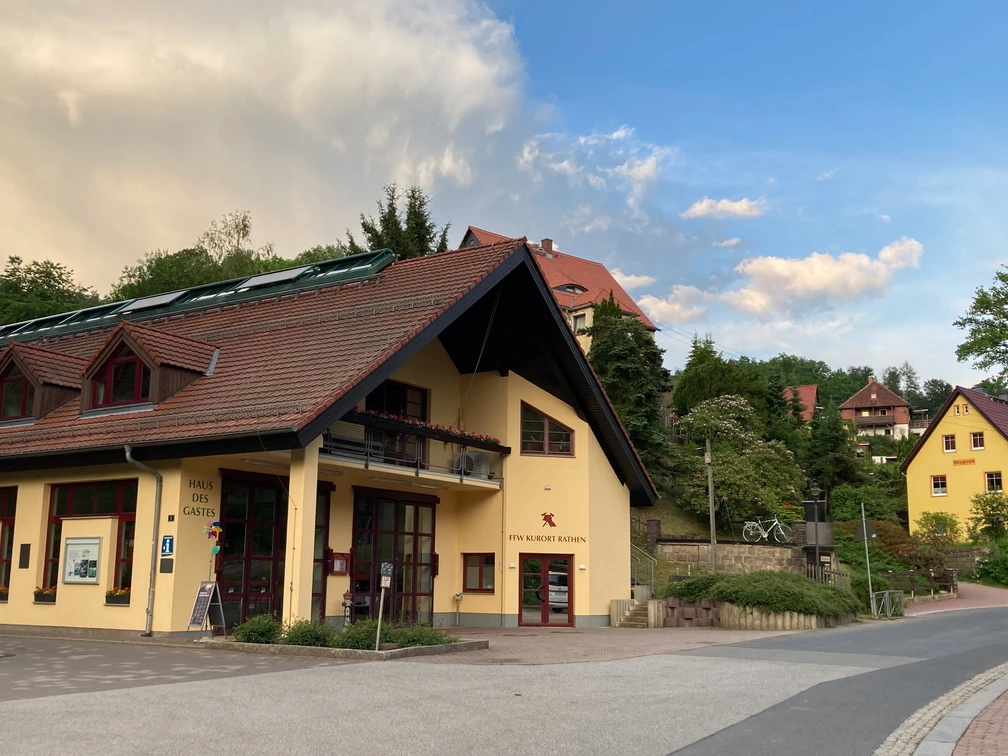 Haus des Gastes außen Gelbes Gebäude mit rotem Dach und Solarpanelen, umgeben von grünen Bäumen und weiteren Häusern, unter blauem Himmel mit Wolken.