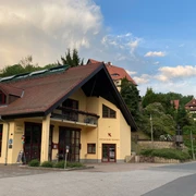 Haus des Gastes außen Gelbes Gebäude mit rotem Dach und Solarpanelen, umgeben von grünen Bäumen und weiteren Häusern, unter blauem Himmel mit Wolken.