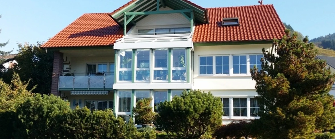 Ein helles zweieinhalbstöckiges Haus mit rotem Dach.. In beiden Stockwerken viele Fenster und ein Wintergarten.