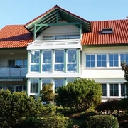 Ein helles zweieinhalbstöckiges Haus mit rotem Dach.. In beiden Stockwerken viele Fenster und ein Wintergarten.