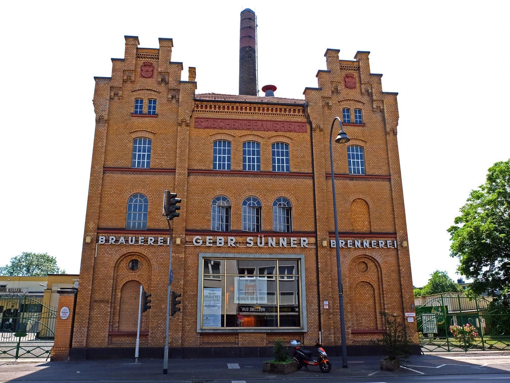 Brauwelt in Cologne-Kalk Historisches Brauereigebäude in Köln-Kalk mit markanter Ziegelfassade und großen Fenstern bei blauem Himmel.Historic brewery building in Cologne-Kalk with striking brick façade and large windows under a blue sky.