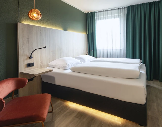 Business Doppelzimmer im ACHAT Hotel Monheim am Rhein Doppelzimmer mit modernem Design, geräumigem Bett, schickem Sessel, Holzdetails und eleganter Beleuchtung.