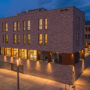 Hotel Neues Pastorat in Heiligenhaus Modernes Backsteingebäude mit großen Fenstern, warm beleuchtet in einer ruhigen Umgebung am Abend.