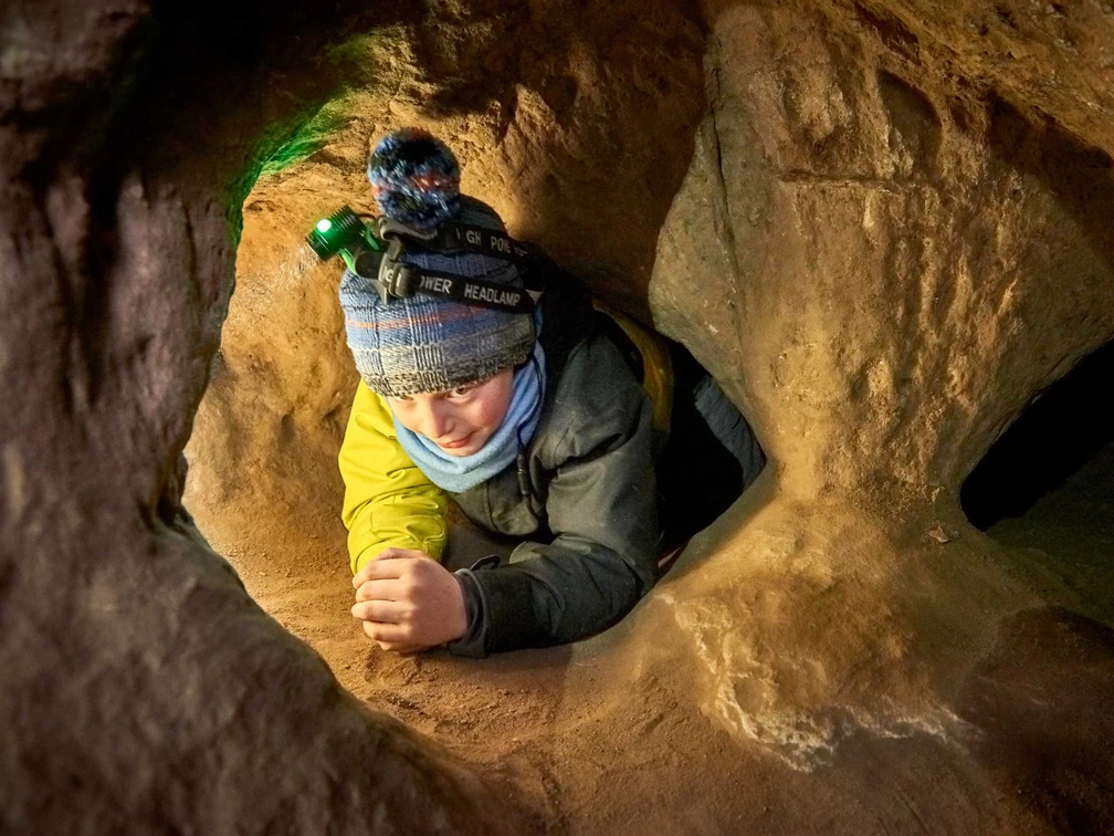 Kleine Höhlenforscher in der Sächsischen Schweiz Kind mit Mütze und Stirnlampe kriecht durch einen engen, sandfarbenen Höhlentunnel. Die Atmosphäre wirkt abenteuerlich und konzentriert.