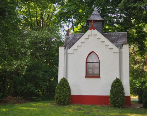 Kleine weiße Kapelle mit rotem Sockel, gotischem Fenster und grüner Umgebung in Hövelhof.