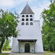Weiße Kirche mit grauem Turm, sechs Bogenfenstern und grünem Umfeld durch Bäume flankiert.