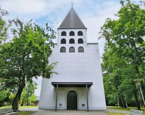 Weiße Kirche mit grauem Turm, sechs Bogenfenstern und grünem Umfeld durch Bäume flankiert.