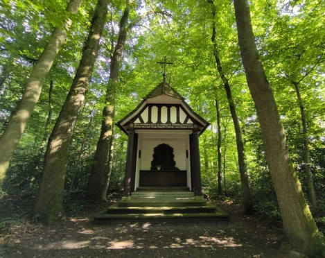Eine kleine, weiße Kapelle mit Fachwerkelementen, umgeben von dichtem, grünem Wald.
