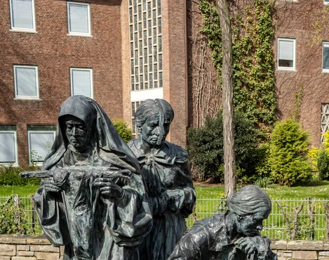 Edith Stein memorial Das Edith-Stein-Denkmal zeigt drei bronzene Frauenfiguren in feierlichen Posen, umgeben von einem gepflasterten Platz und Grünflächen.The Edith Stein monument shows three bronze female figures in solemn poses, surrounded by a paved square and green areas.