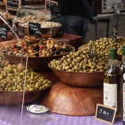 Feinkost Kaya (c) Pixabay Auf einem Tisch mit Tischtuch stehen rechts verschiedene Flaschen von Olivenöl und links daneben mehrere Holzschalen gefüllt mit jeweils verschiedenen Olivensorten.