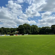 Sportplatz Gristede.jpg Weitläufiger, grün gehaltener Fußballplatz unter blauem Himmel mit Wolken, umgeben von Bäumen.