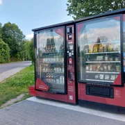 Proviantomat in Ostrau Roter Verkaufsautomat mit Snacks und Getränken, aufgestellt neben einer Landstraße, umgeben von grünen Bäumen und Wiese unter blauem Himmel.