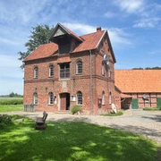 Noltesche Mühle Bruchhausen-Vilsen (2).jpg Historische Backsteinmühle mit zwei Etagen, umgeben von grünem Rasen, Bäumen und einem klaren Himmel.