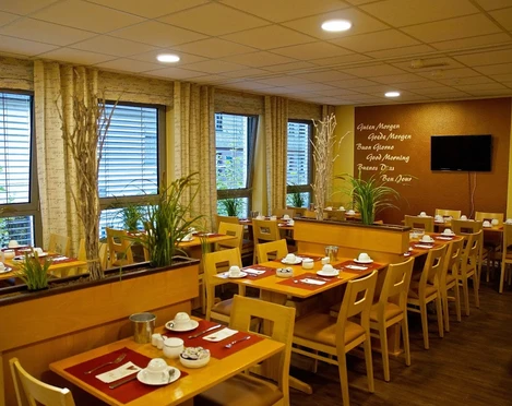 Frühstücksraum im Hotel Alberga in Mettmann Heller Frühstücksraum mit Holzmöbeln, eingedeckt für Gäste, bietet Ausblick durch große Fenster.