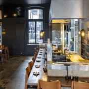 Daikan Izakaya Bar Innenansicht vom Restaurant mit dunklen Wänden und hellen Holzmöbeln. Hinter der Theke, hinter Glas die Küche.Interior view of the restaurant with dark walls and light wooden furniture. Behind the bar, behind glass, the kitchen.