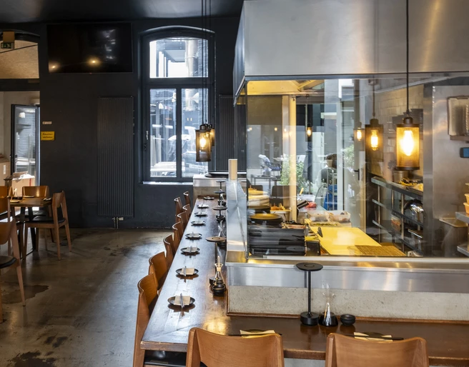 Daikan Izakaya Bar Innenansicht vom Restaurant mit dunklen Wänden und hellen Holzmöbeln. Hinter der Theke, hinter Glas die Küche.Interior view of the restaurant with dark walls and light wooden furniture. Behind the bar, behind glass, the kitchen.