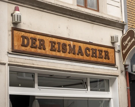 Der Eismacher Fassade eines Geschäfts mit dem Holzschild „Der Eismacher“ über der Eingangstür.Façade of a store with the wooden sign "Der Eismacher" above the entrance door.