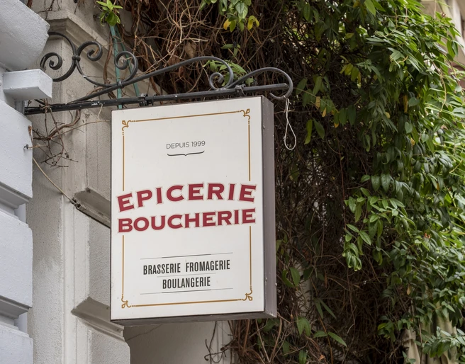 Epicerie Boucherie Ein Schild befestigt an einer verzierten Metallstange schmückt die Fassade eines Altbaus. Umgeben von grünem Gewächs. A sign attached to an ornate metal pole decorates the facade of an old building. Surrounded by green plants.