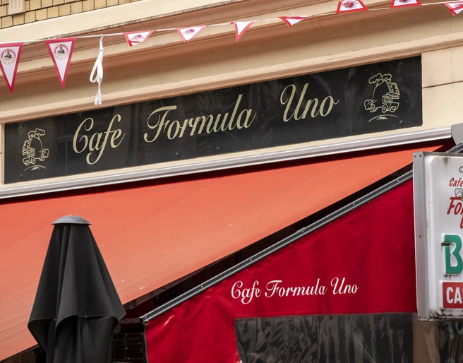 Formula Uno An der Fassade hängt ein schwarzes Schild mit der Aufschrift Formula Uno. Eine Wimpelgirlande flattert im Wind.A black sign with the inscription Formula Uno hangs on the façade. A garland of pennants flutters in the wind.