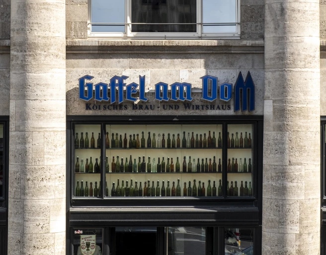 Gaffel am Dom Außenansicht auf das Brauhaus. Zwei markante Säulen rahmen ein Fenster ein, das mit bunten Flaschen dekoriert ist.Exterior view of the brewery. Two prominent columns frame a window decorated with colorful bottles.
