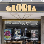 Gloria  Schwarzes Schild mit goldener Aufschrift "GLORIA" über einem Kinoeingang mit bunten Filmplakaten.Black sign with gold lettering "GLORIA" above a cinema entrance with colorful movie posters.