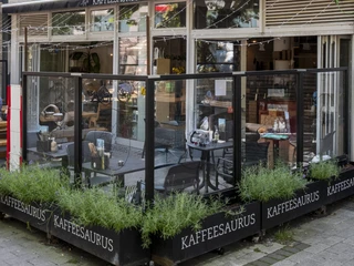 Kaffeesaurus Ansicht auf die Fassade des Café mit Außengastronomie. Ein bestuhlter Bereich ist durch eine transparente Wand vom Gehweg abgetrennt. View of the café façade with outdoor seating. A seating area is separated from the sidewalk by a transparent wall.