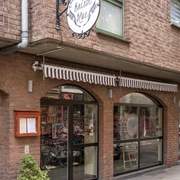 Maison Blue Ansicht auf die Hausfassade mit bodenlanger Fensterfront. Die Fenster sind in verschnörkelter Schrift beschrieben und eine eingefahrene gestreifte Markise befindet sich direkt darüber. View of the house facade with floor-to-ceiling windows. The windows are decorated with ornate lettering and a rolled-up striped awning is located directly above them.