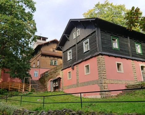 Baude Großer Winterberg Zwei traditionelle Häuser mit rosa und schwarzer Fassade stehen auf einem grünen Hügel, umgeben von Bäumen unter einem bewölkten Himmel.
