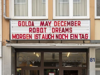 Odeon  Das Bild zeigt den Eingang des Odeon Kinos mit einem Vorbau aus Glas. Über der Tür hängen Filmankündigungen in großen roten Buchstaben auf einer weißen Anzeigetafel. Ein Zugangsweg mit sanfter Rampe im Vordergrund verstärkt die Barrierefreiheit für alle Besucher*innen.The picture shows the entrance to the Odeon movie theater with a glass porch. Above the door, movie announcements hang in large red letters on a white display board. An access path with a gentle ramp in the foreground reinforces the accessibility for all visitors.