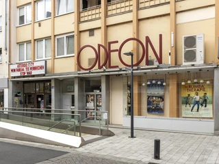 Odeon  Das Bild zeigt die Fassade eines Kinos namens "Odeon" in einer urbanen Umgebung. Große rote Buchstaben bilden den Namen auf einem Vordach. Im Schaukasten sind Filmplakate zu sehen, die aktuelle Filme ankündigen. Angrenzende Gebäudeteile und eine Gehwegführung aus Glas bieten Barrierefreiheit.The picture shows the façade of a cinema called "Odeon" in an urban environment. Large red letters form the name on a canopy. Film posters announcing current movies can be seen in the showcase. Adjacent parts of the building and a glass walkway provide accessibility.