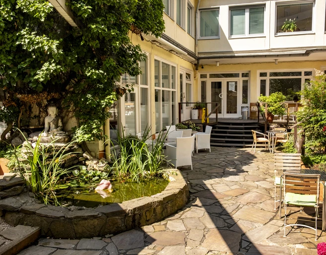 Oshos Place Ein ruhiger Innenhof mit einem kleinen, von Pflanzen umgebenen Teich. Sitzmöglichkeiten im Freien laden zum Verweilen ein, während üppiges Grün den Charme des Ortes unterstreicht.A quiet courtyard with a small pond surrounded by plants. Outdoor seating invites you to linger, while lush greenery emphasises the charm of the place.