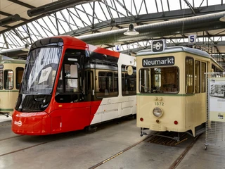 Thielenbruch Tram Museum Drei historische Straßenbahnen stehen in einer Halle nebeneinander: zwei ältere Modelle mit creme- und grüner Lackierung sowie eine moderne rote Straßenbahn. Schilder über den Fahrzeugen weisen auf Haltestellen wie "Neumarkt" hin. Die lichte Halle ist aus Stahl und Glas gefertigt.Three historic streetcars stand side by side in a hall: two older models with cream and green paintwork and a modern red streetcar. Signs above the vehicles indicate stops such as "Neumarkt". The light-filled hall is made of steel and glass.